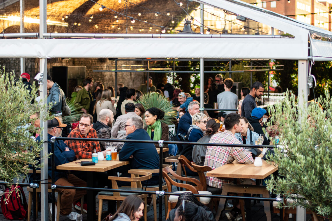 A photo of people under the Courtyard's covered space, seated at tables, enjoying beer in cans and glasses.