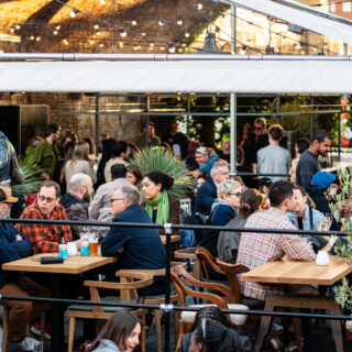 A photo of people under the Courtyard's covered space, seated at tables, enjoying beer in cans and glasses.