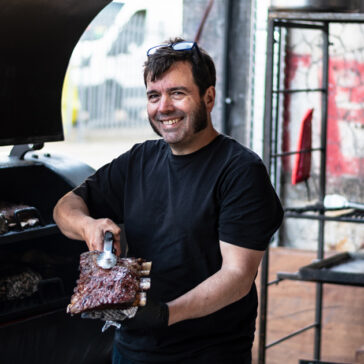Martin from From The Ashes holds up some beautifully smoked ribs to the camera with a pair of tongs in front of the smoker.