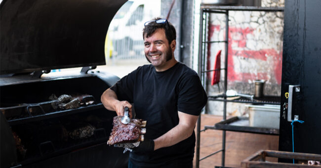 Martin from From The Ashes holds up some beautifully smoked ribs to the camera with a pair of tongs in front of the smoker.