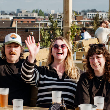Will, Jess and Hannah of The Five Points Sales Team are sat at a table at Netil 360. Jess has her hand up. There are cups in front of them with beer.