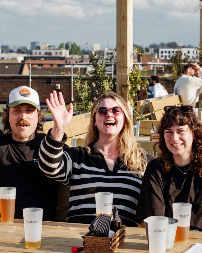 Will, Jess and Hannah of The Five Points Sales Team are sat at a table at Netil 360. Jess has her hand up. There are cups in front of them with beer.