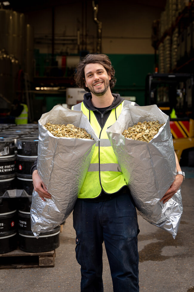 Max holds up two silver bags full of hops and he is smiling. He is wearing a hi-vis yellow vest.