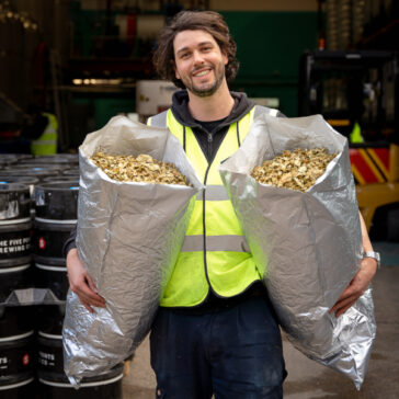 Max holds up two silver bags full of hops and he is smiling. He is wearing a hi-vis yellow vest.